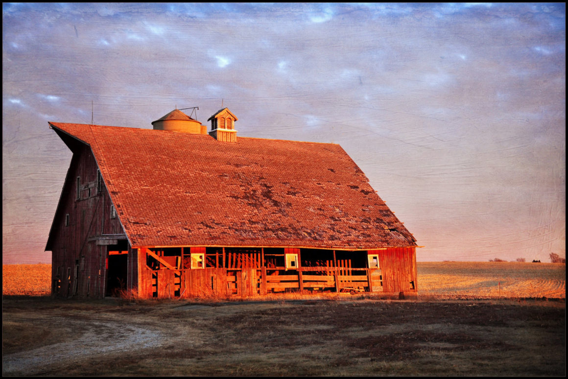 an abandoned barn in the middle of a field, at sunset